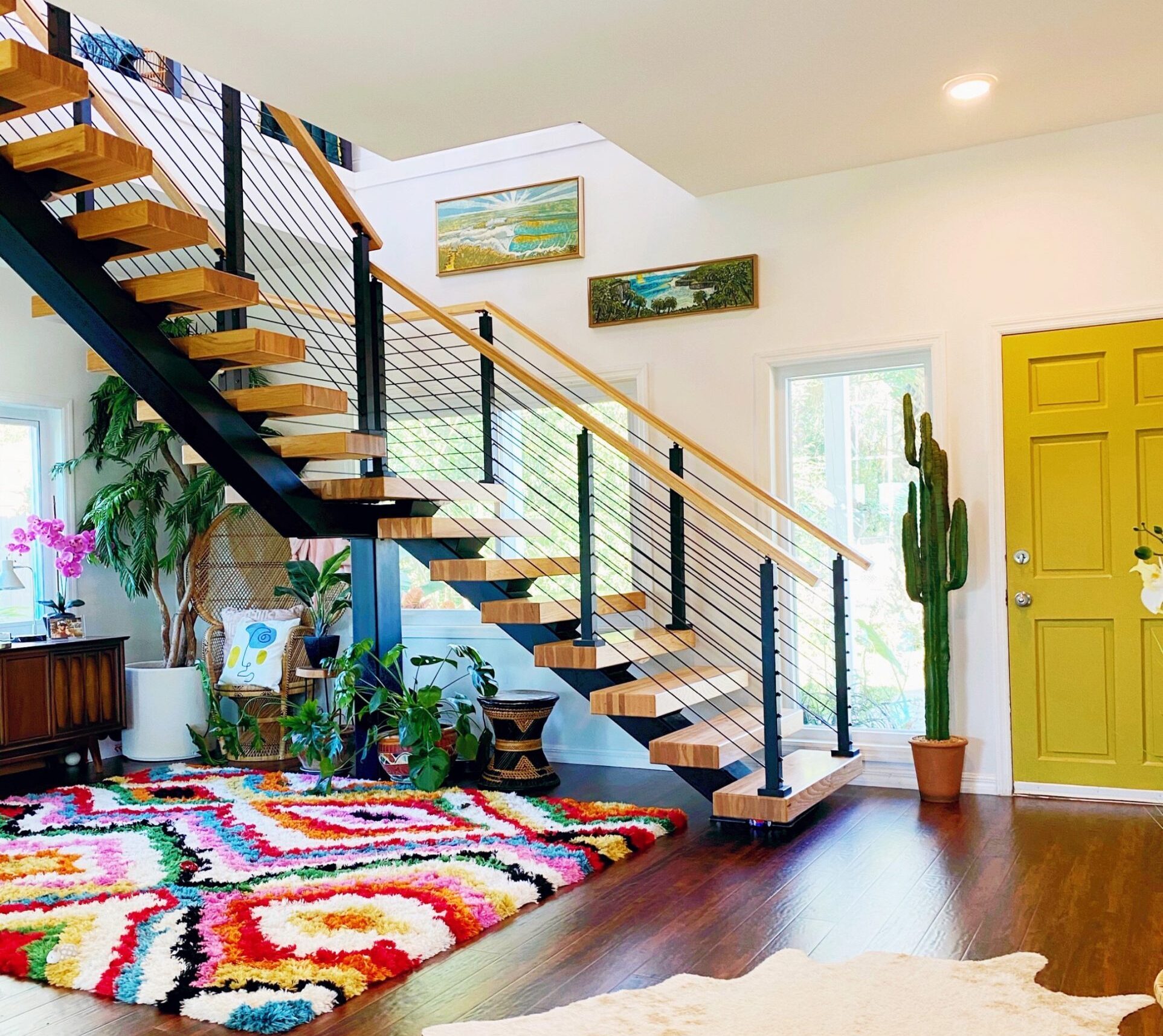 This fun home has a yellow door with a cactus next to it. There is a bright, multi-colored rug in the center of the room with various plants under the floating staircase. 